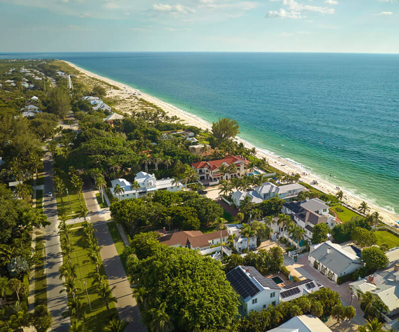 Aerial view of a coastal neighborhood in Tampa Bay, Florida, with modern houses enveloped by lush greenery. The homes are near a sandy beach and turquoise ocean, while streets lined with palm trees run parallel to the shoreline.