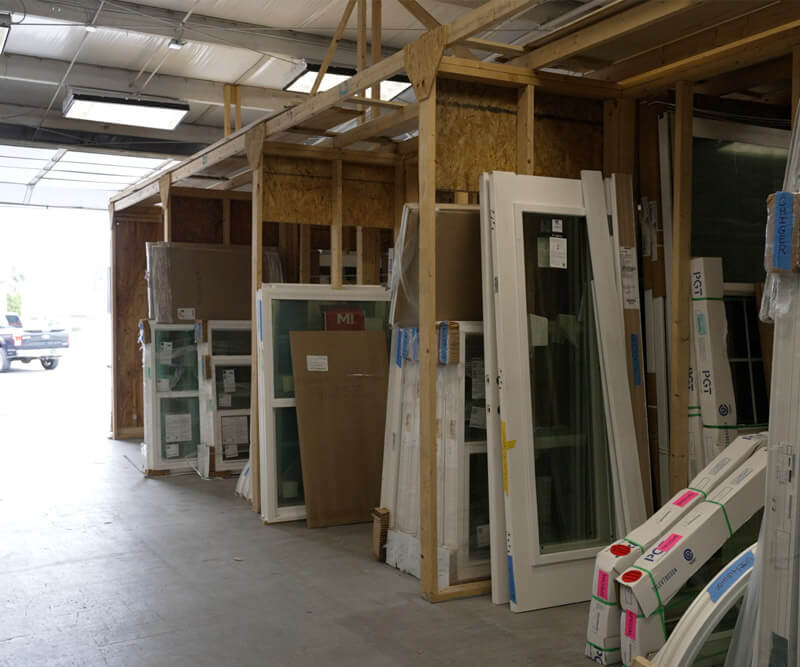 A storage area with factory-made windows and doors leaning against a wall. Several items are packaged in cardboard and plastic. Through the open entrance, a red vehicle is visible outside.