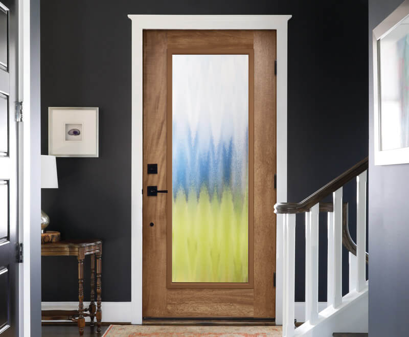 A woodgrain fiberglass door with a Chinchilla glass panel featuring blue, white, and green hues is set in a dark-painted entryway. To the left, a small framed artwork adorns the wall above a wooden table with a lamp. A white staircase railing elegantly graces the right side.