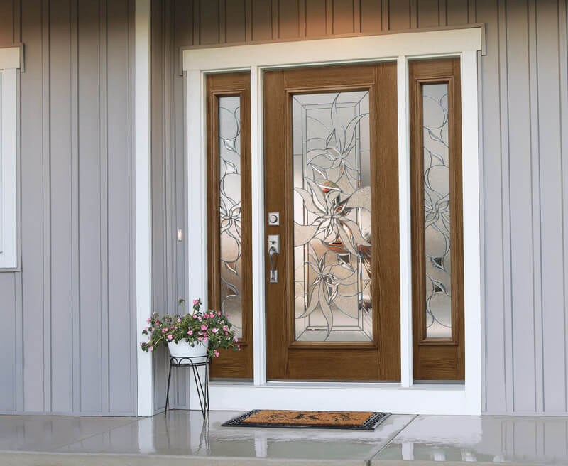 Front door with decorative frosted glass, showcasing a floral design. Flanked by narrow sidelights featuring matching patterns. A small flower pot with pink blooms sits on a stand next to the entrance, complemented by a doormat on the concrete step.