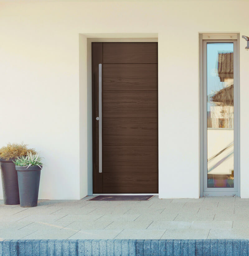 A modern house entrance features a woodgrain fiberglass door with a narrow vertical window beside it. Two tall planters with assorted plants stand gracefully on the left, complementing the sleek design. Light-colored tiles pave the area, adding an elegant touch to the setting.