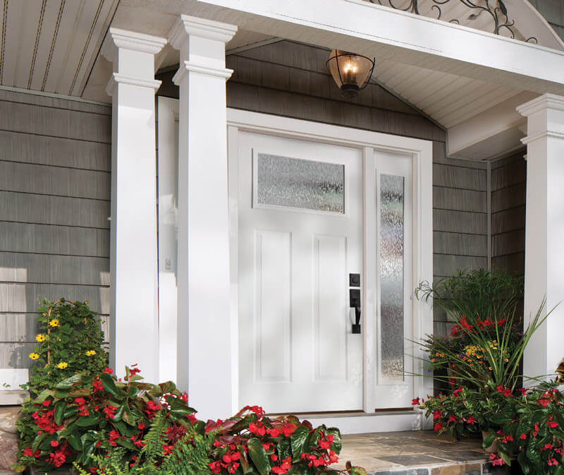 A white front door with streamed privacy glass panels is flanked by two tall white columns. Red flowers and green plants adorn garden beds on each side of the entrance. A black lantern elegantly hangs from the ceiling above, casting a warm glow over the inviting space.