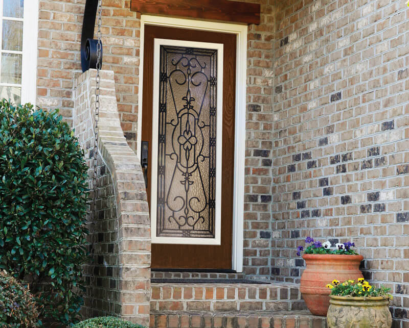 A decorative woodgrain front door with intricate wrought iron design is set in a brick wall. A curved brick wall leads to the porch. A terracotta pot with colorful flowers is on the right, and a large bush is on the left.