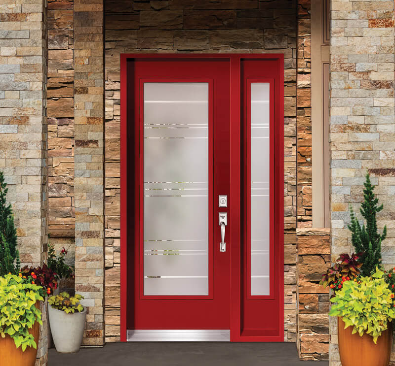 A red entry door with sandblasted privacy glass panels, framed by stone walls. Potted plants with green and yellow foliage are placed on either side, enhancing the entrances welcoming appearance.