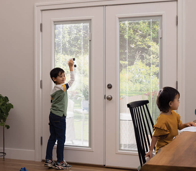 Two children are indoors by the double entry doors with blinds. The boy, on his toes, adjusts a handle on the door. Meanwhile, the girl in a mustard top sits at a table. A leafy view is visible outside through the doors.