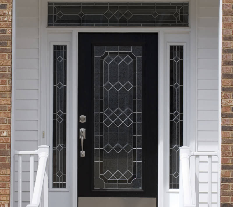 A black entry door system with ornate geometric glass panels is framed by sidelights and a transom window featuring matching designs. The door is flanked by white siding and red brick walls, with white railings on each side of the entrance.