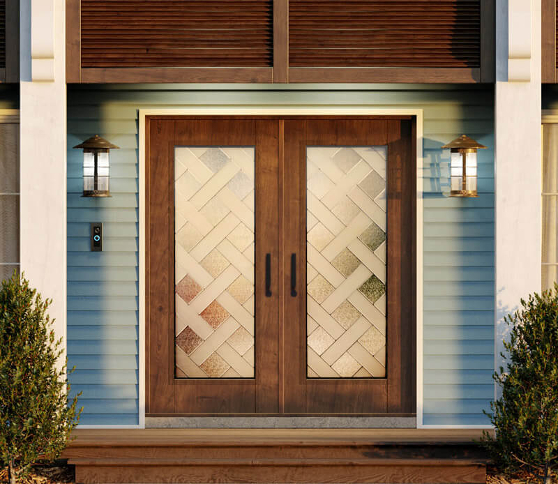 A set of stunning double front doors in woodgrain fiberglass, adorned with frosted glass panels featuring a crisscross pattern. Flanked by sleek wall lanterns, the entrance is surrounded by blue siding and small green bushes on either side.