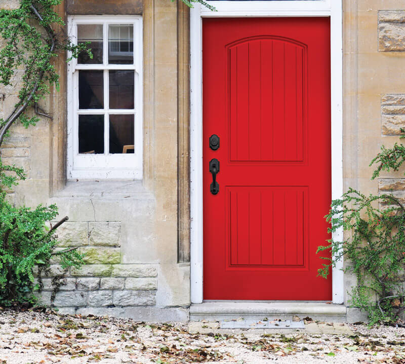 A vibrant red, smooth-grained solid fiberglass door set in a stone facade, next to a window with white frames. The area is adorned with green climbing plants, and the ground is covered with dry leaves. This door features a black handle and a keyhole.