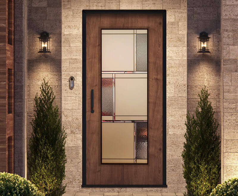 A fiberglass door with a decorative glass pane featuring geometric patterns. The doorway is flanked by two small shrubs and illuminated by two wall-mounted lanterns, creating a welcoming entrance on a stone wall background.