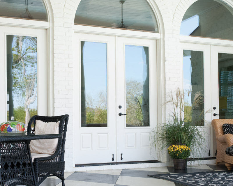 A white brick patio showcases double front doors in glass, with wicker furniture and potted plants adding charm. A black wicker chair and table sit on the left, while a beige cushioned chair is on the right. Bright sunlight bathes the scene in warmth.