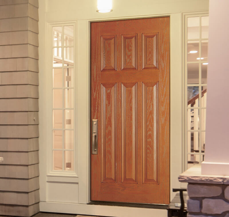 A 6-paneled, Craftsman-style fiberglass door stands prominently with a classic design, flanked by tall, narrow windows. The warmly lit interior provides an inviting backdrop, while a hint of a stone pillar is glimpsed to the right.