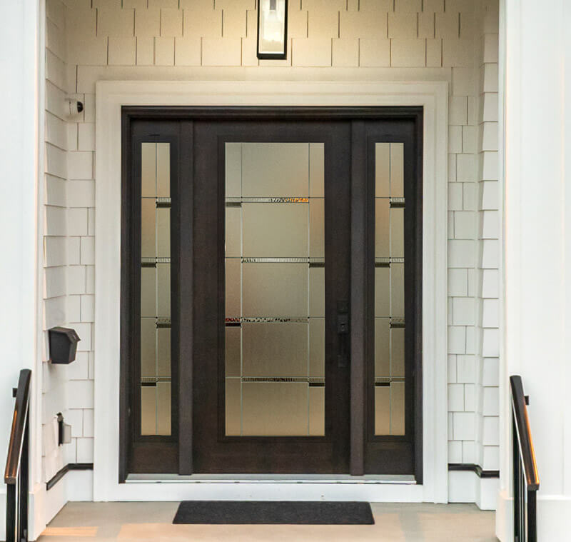 A modern dark-stained woodgrain fiberglass door with frosted glass panels and decorative horizontal lines. The entrance is flanked by white siding and illuminated by a rectangular light fixture above, with a welcome mat lying in front.