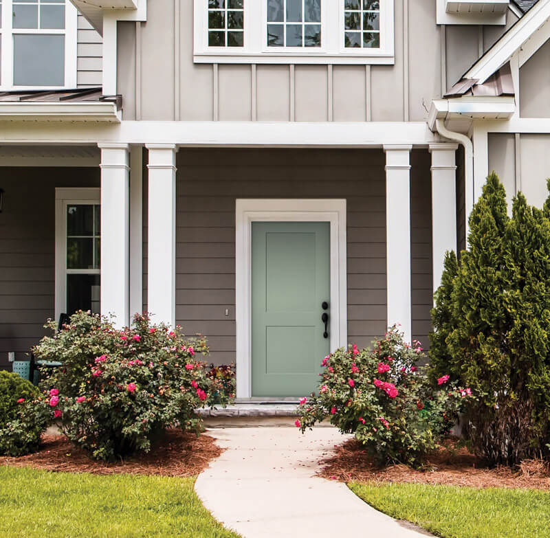 Front view of a house showcasing a gray facade with a light green front entry door. The entrance is framed by white pillars and surrounded by blooming rose bushes. A concrete walkway leads to the porch, with neatly trimmed grass in the foreground.