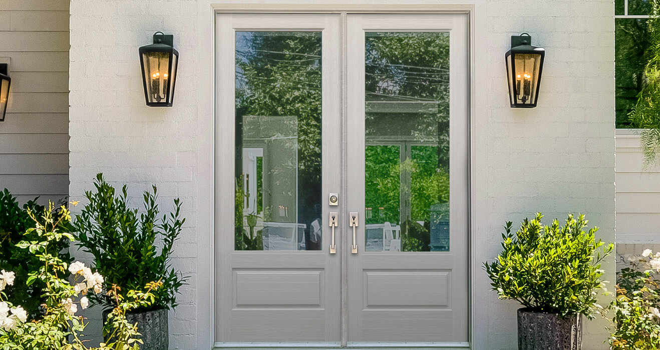 The entry door, featuring sleek white frames and 3/4 glass, stands at the center of the image. Flanked by elegant black wall lanterns and vibrant potted greenery, it reflects a lush garden scene with trees and plants, suggesting a sunny day.