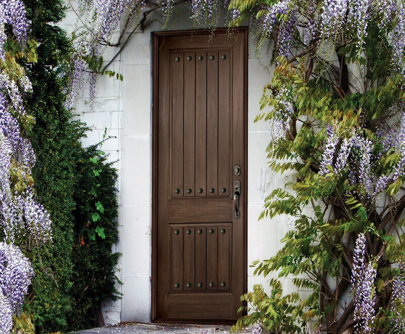 A woodgrain fiberglass door with metal studs is framed by lush green foliage and cascading purple wisteria flowers, set against a white stone wall.