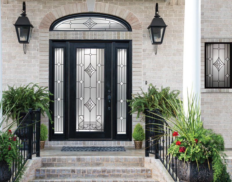 A grand entrance with a black entry door featuring intricate glass designs and double sidelites. Flanked by two large lanterns and lush ferns, the brick steps lead up to the doorway. A symmetrical design with a window of matching style is on the right.