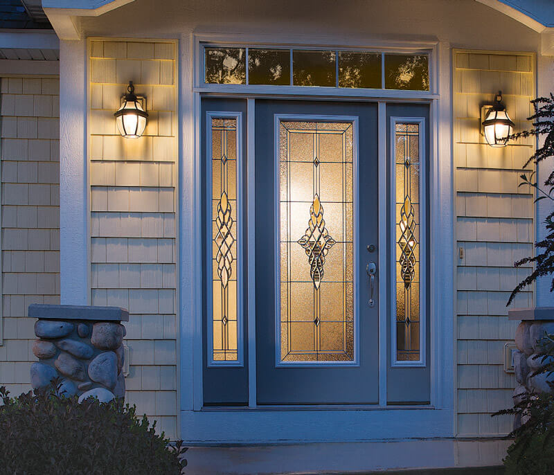 A front entry door painted a vibrant blue, featuring decorative glass panels and flanked by two ornate sidelights, captures Floridas sunny charm. Two wall-mounted lanterns illuminate the entrance, complemented by beige siding and stone accents adorning the house exterior.