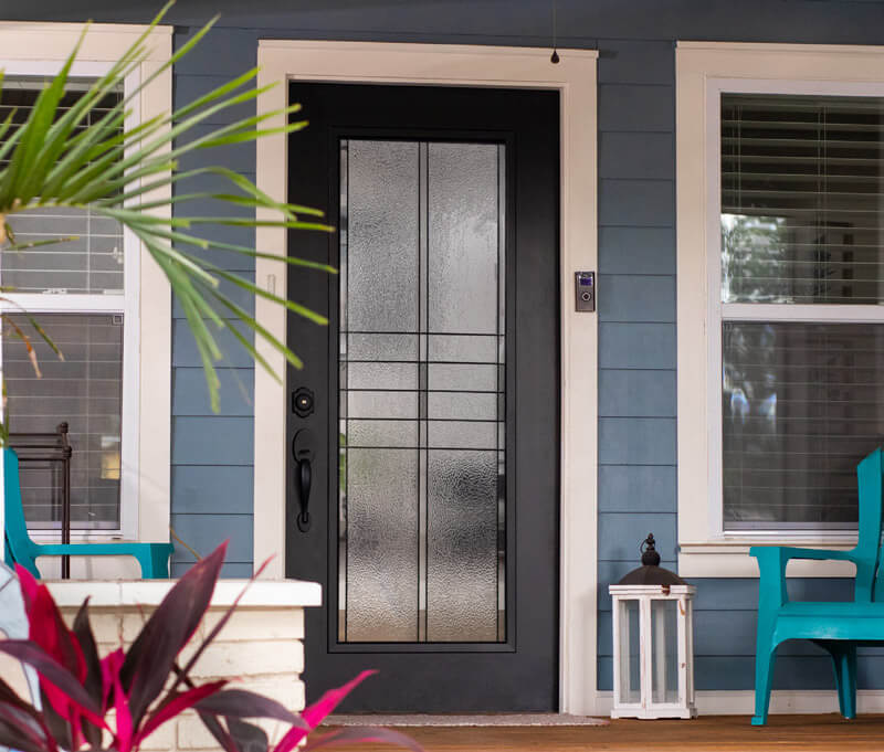 A modern porch showcases a front door with decorative glass panels, flanked by windows with blinds. Blue siding enhances the exterior. Two teal chairs and a white lantern grace the porch, while foliage peeks into view.