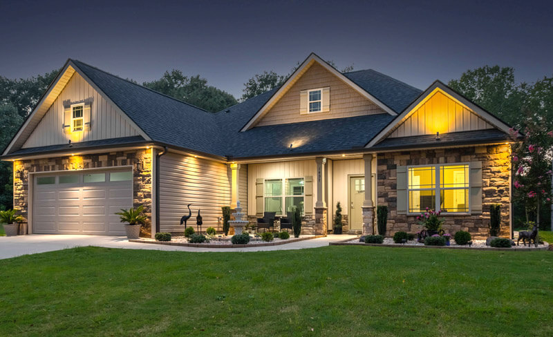 Single-story house with a peaked roof, lit warmly against a twilight sky. Features a two-car garage, stone and siding exterior, and neatly maintained lawn. Decorative plants and statues adorn the front porch area.