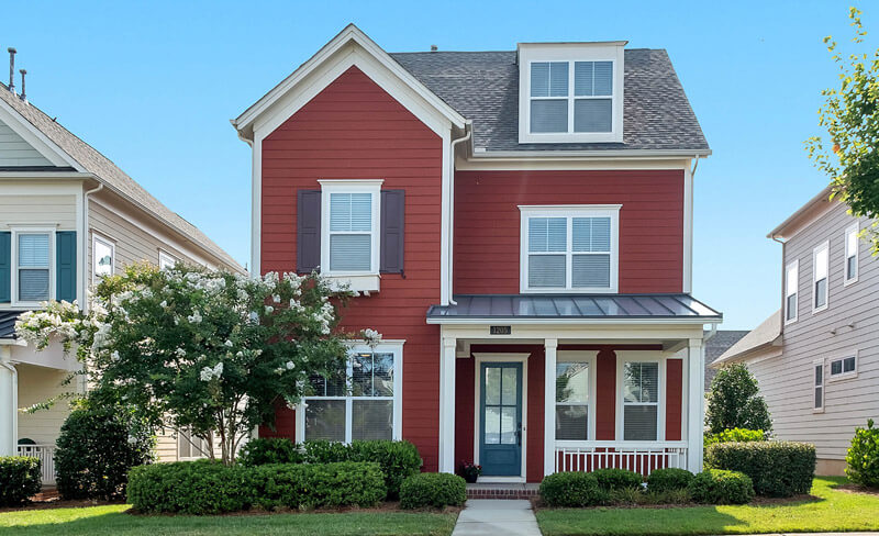 A two-story red house with a charming front porch and inviting front door boasts white trim, dark shutters, and a manicured lawn. Two trees elegantly flank the entrance as the house sits gracefully between similar homes under a clear blue sky.