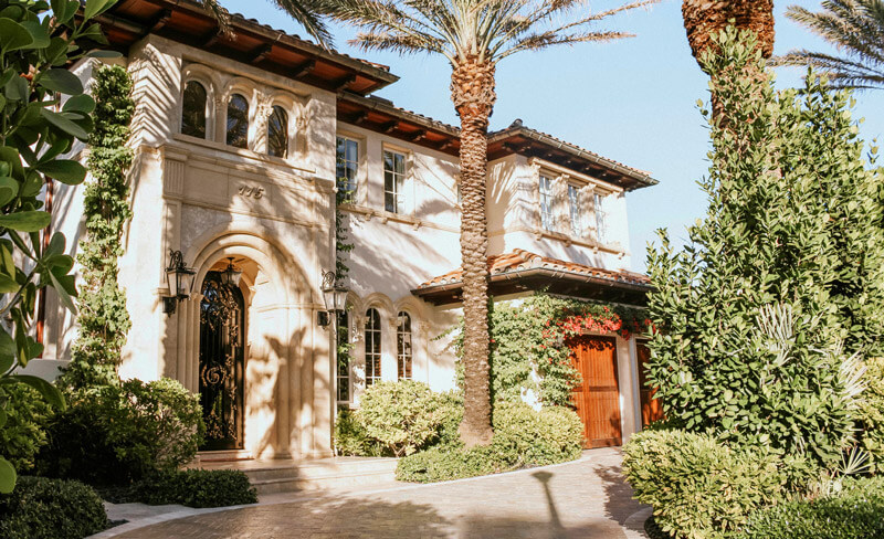 Mediterranean-style house with arched windows and a charming wrought-iron, impact-resistant front entry door, surrounded by lush greenery and tall palm trees. The light-colored facade features intricate detailing, with a sunny driveway leading to a wooden garage door.