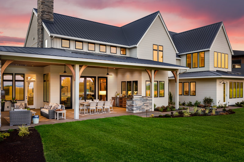 Spacious modern farmhouse with large windows and a black metal roof amidst a sunset sky. Garden doors open to a patio featuring outdoor furniture and a dining set, overlooking the lush lawn and garden. The warm interior lights create an inviting atmosphere.