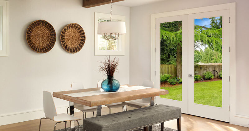 Modern dining room with a wooden table, white chairs, and a gray bench. A blue vase with twigs sits on the table. Round wicker wall decor hangs beside garden doors with clear glass that overlook a green backyard with trees. White walls and light wood flooring.