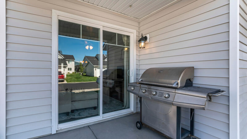 A backyard patio showcases a stainless steel gas grill beside sleek patio doors. The light gray siding complements the setup, and a small outdoor light is mounted above the grill. Reflections in the doors reveal a lush green lawn and nearby houses.