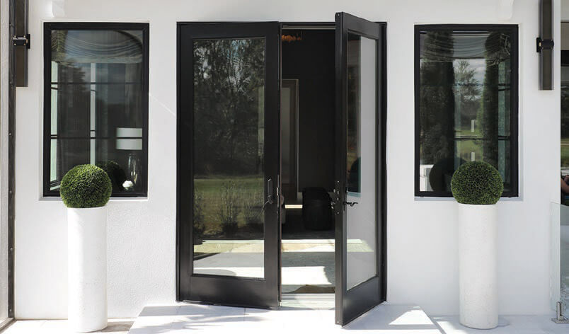 A modern entrance showcases PGT WinGuard French Doors, their sleek black frames cradling the glass, one slightly ajar. Flanking the doors are two white pedestals topped with round green shrubs against a white facade punctuated by rectangular windows.