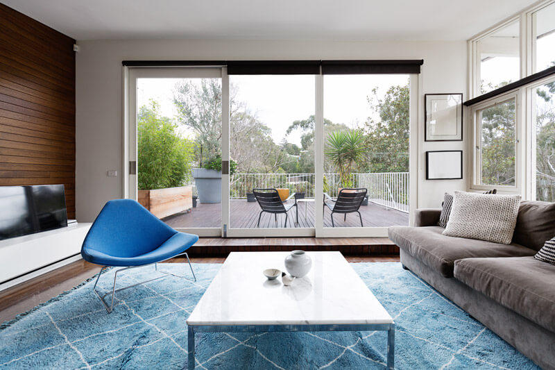 A modern living room with patio doors offers a stunning view of the deck and trees. It features a blue armchair, gray sofa, and white marble coffee table on a blue rug. The flat-screen TV complements the serene setting, while the deck, equipped with chairs and a table, is surrounded by lush greenery.