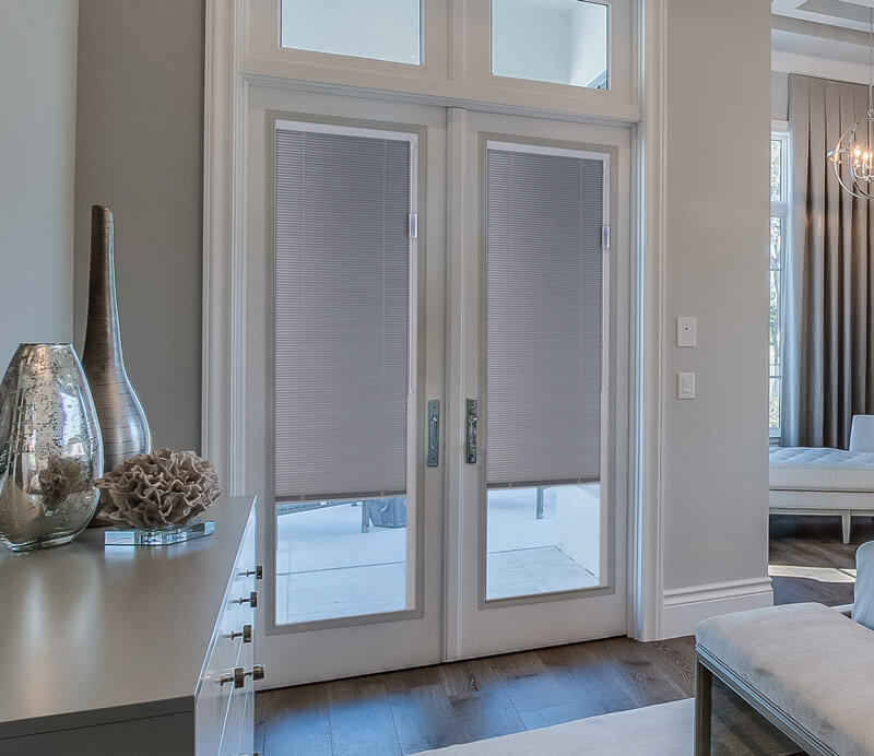 A modern living room features French doors adorned with pleated blinds. The space boasts light gray walls, a white ceiling, and dark hardwood floors. On the left, a sideboard displays decorative vases, while part of a sofa is visible on the right.