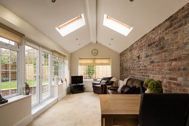 A cozy sunroom with large windows, garden doors, and skylights, featuring a brick accent wall. The room has two brown leather sofas, a small TV, and a wooden table with a black chair. Outside, a fence encloses a grassy yard. A wall clock is above the window.