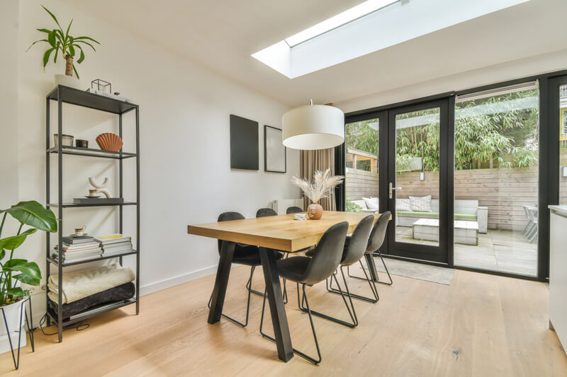 Modern dining room with a wooden table and six black chairs. A potted plant and shelves with books adorn the left, while patio doors reveal an inviting outdoor space. A large, round white pendant light elegantly hangs above, illuminating the harmony between indoor charm and outdoor tranquility.