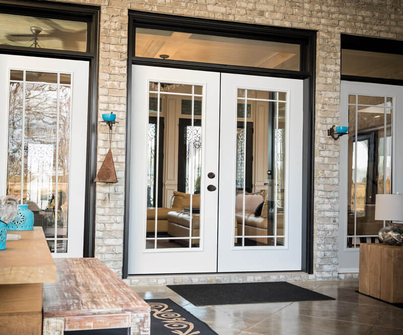 A pair of patio doors with glass panels and white frames set in a stone brick wall. The interior reveals a cozy couch and decor. A wooden bench sits to the left, while a doormat is placed invitingly in front of the doors.