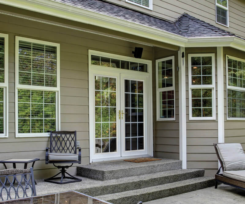 A patio with steps leads to garden doors with grilles on a beige house. Outdoor seating, including a chair and cushioned bench, is set against large windows with white frames. Trees are beautifully reflected in the glass.