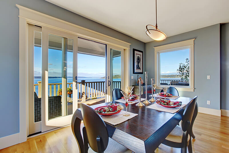 Dining room with a wooden table set for four, red plates, and decorative wine glasses. A large window and sliding patio doors reveal a balcony view of the ocean and blue sky. Walls are light blue with white trim, and a modern pendant light hangs above.