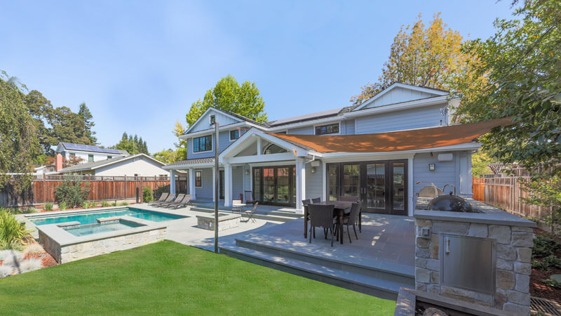 A modern two-story house with light gray siding features elegant garden doors leading to a backyard patio with a dining table, stone grill area, and canopy overhang. A rectangular pool and hot tub are surrounded by a lush green lawn and landscaped plants, framed by trees.