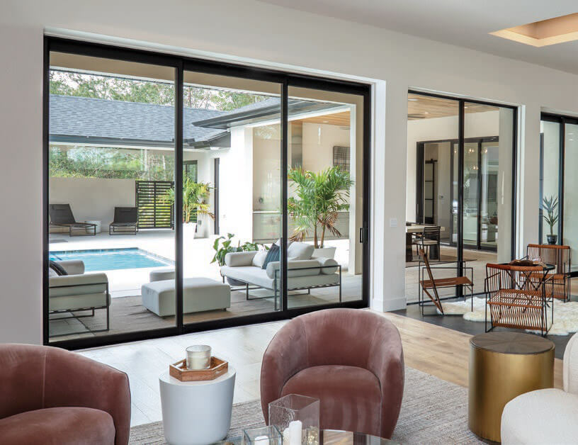 Modern living room with plush chairs and tables, centered on expansive patio doors that open to an outdoor seating area by the pool. Surrounded by lush greenery, natural light floods in, creating a seamless indoor-outdoor transition.