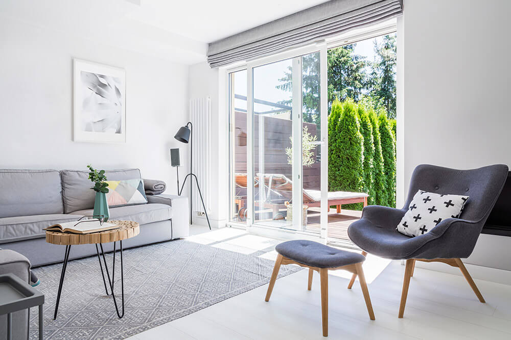 A bright, modern living room with a large window and sliding glass doors leads to a patio adorned with green shrubs. It features a gray sofa, a matching armchair with footrest, a wooden side table, and a floor lamp. A framed abstract art piece graces the wall.