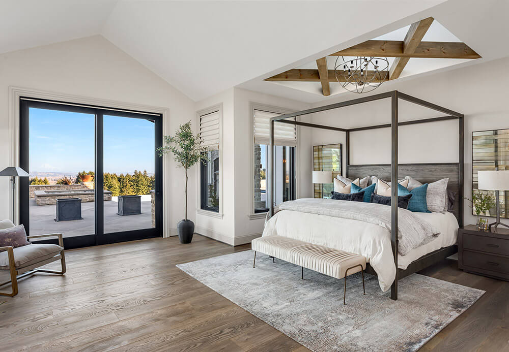 A spacious bedroom with a large canopy bed, light bedding, and a cushioned bench at the foot. Soft rug on wooden floors complements black patio doors leading to an outdoor terrace. Floor-to-ceiling windows and wooden beams enhance the modern light fixture above.