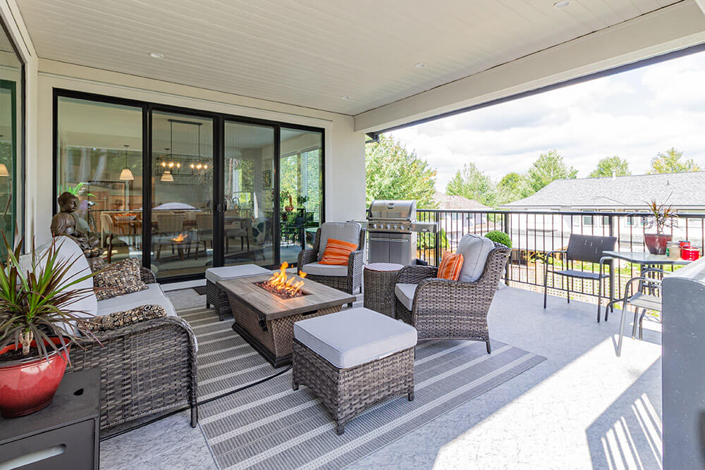 Covered patio with wicker furniture, cushions, and a central fire table. A rug defines the seating area, and a grill is positioned near the black sliding patio doors. Trees and rooftops are visible in the background under a partly cloudy sky.