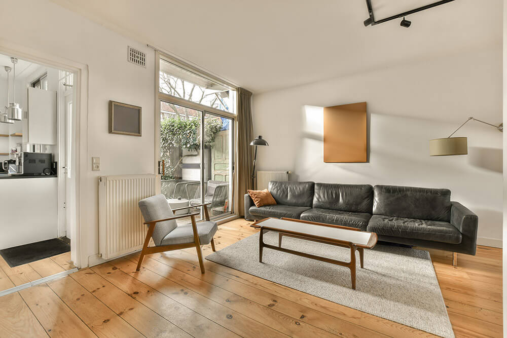 A modern living room features a black sofa, wooden coffee table, and gray armchair on a light rug. Sliding glass doors offer a view of greenery, complemented by a standing lamp and wall art. A doorway leads to a kitchen with white cabinets, while wooden floors enhance the light walls.