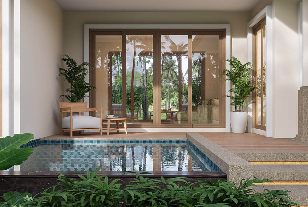 A modern patio with a small pool, surrounded by lush greenery. Wooden chairs and tables sit on the deck, with large glass patio doors leading inside. Two potted plants are beside the entrance, enhancing the serene, tropical ambiance.