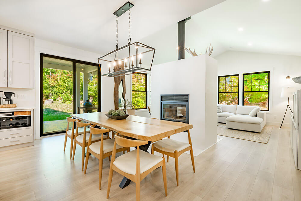 Modern open-concept dining and living area with light wood flooring. A wooden dining table with six chairs sits under a geometric chandelier. Black patio doors lead to a serene outdoor space, while a fireplace divides the dining area from a bright living room with a white sectional sofa and large windows.