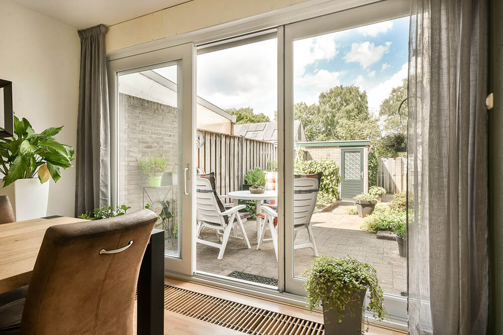 A bright room with patio doors opens to a Florida-style outdoor space. The patio features a table with four white chairs, surrounded by lush greenery and plants. Indoor plants near the door add a touch of nature to the interior. The sky is partly cloudy.