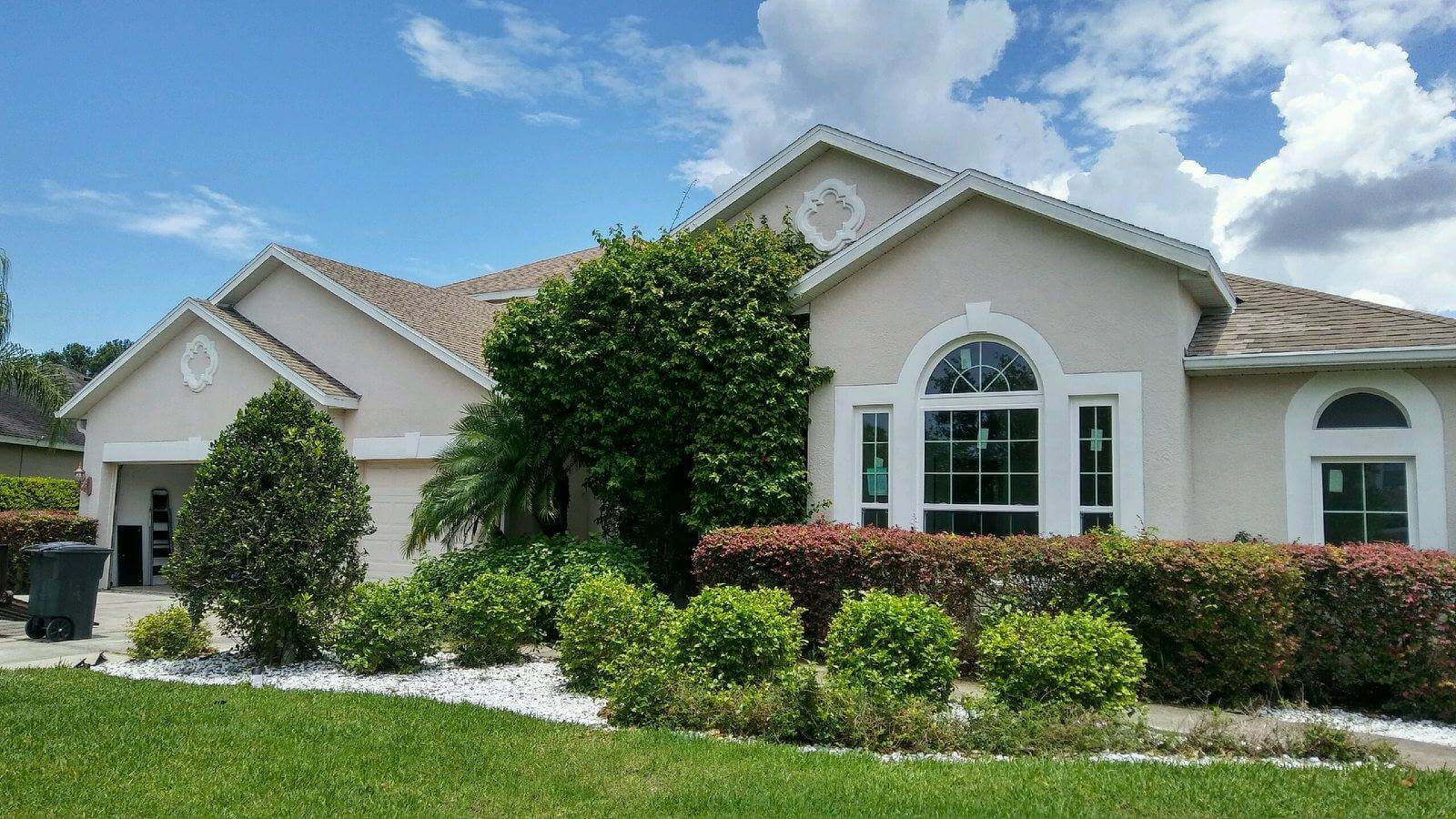 A light-colored single-story house with large arched windows, a two-car garage, and a shingled roof. The front yard is landscaped with bushes, a palm tree, and a small gravel area. The sky is partly cloudy.