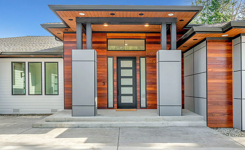 Modern house entrance featuring a mix of wooden and metal paneling. The impact-resistant door is flanked by large square columns and a wide overhang. A row of windows is on the left, and the concrete pathway leads to the front door.