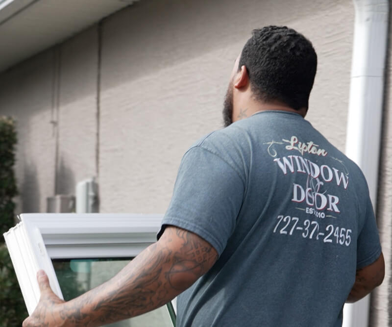 A man carrying a window frame approaches a house. His blue T-shirt displays Lipton Window and Door installer, complete with a contact number. The home boasts a light-colored stucco exterior, providing the perfect backdrop for his task.