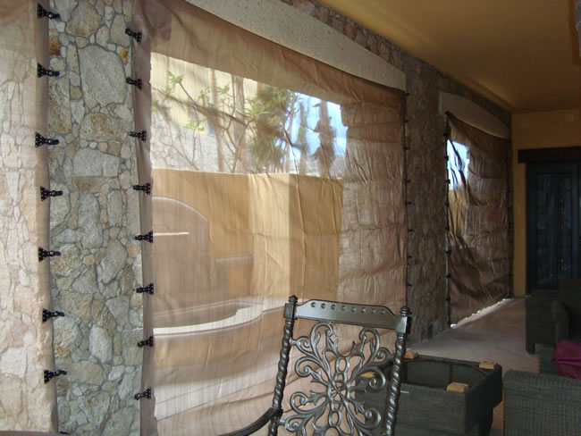 A patio with stone walls features large, fabric storm panels offering hurricane protection for the windows. An ornate metal chair graces the foreground. The space is elegantly furnished with outdoor seating and surrounded by a serene ambiance.