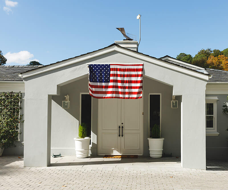 A modern house with a gray exterior and small porch proudly showcases a U.S. flag above the white double front replacement doors in Tampa Bay. Two large potted plants adorn each side of the entrance, framed by a clear blue sky in the background.